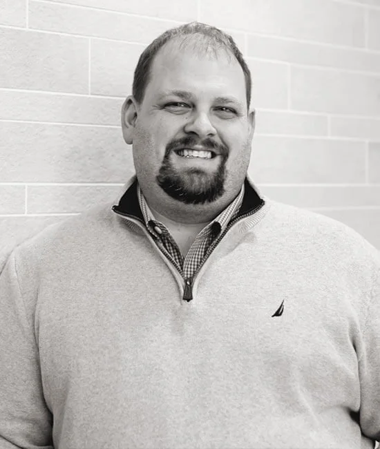 Black and white photo of a man smiling, standing in front of a brick wall, wearing a light-colored quarter-zip sweater over a collared shirt.