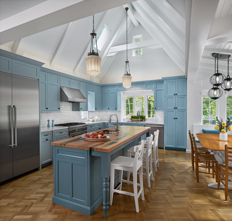 A bright kitchen with a blue island and cabinets, white subway tile backsplash, stainless steel refrigerator and range, wooden dining table with chairs, and large windows letting in natural light.