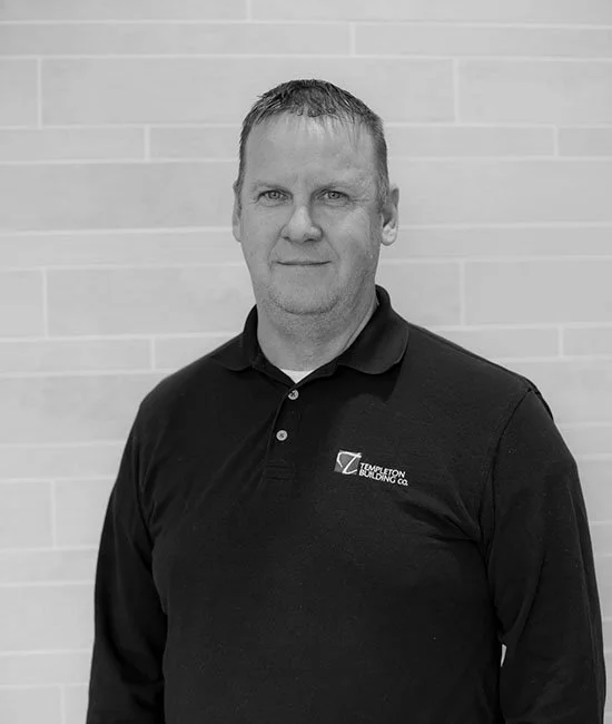 A man with short hair wearing a black collared shirt with a logo standing against a brick wall.