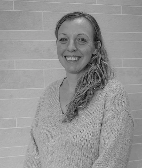 Black and white photo of a smiling woman with long, wavy hair standing in front of a tiled wall.