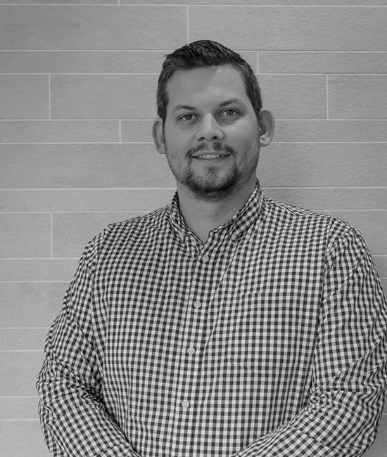 A man with short dark hair and a goatee, wearing a checkered button-up shirt, standing in front of a textured wall.