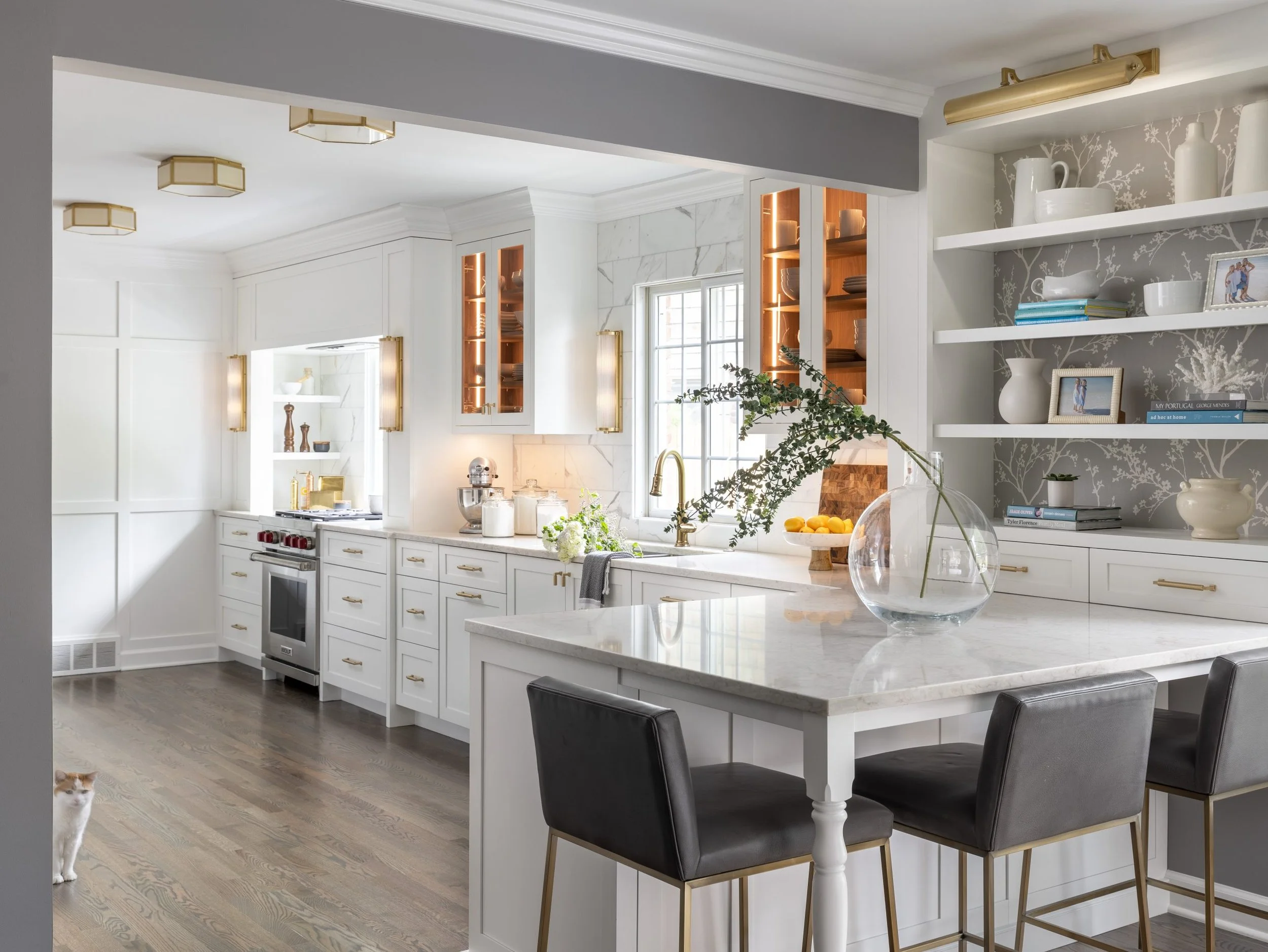 Modern kitchen with white cabinets, marble countertops, open shelving with white dishes and books, gray and white wallpaper, black chairs at a white island with a glass vase containing green foliage, and a cat in the lower left corner.