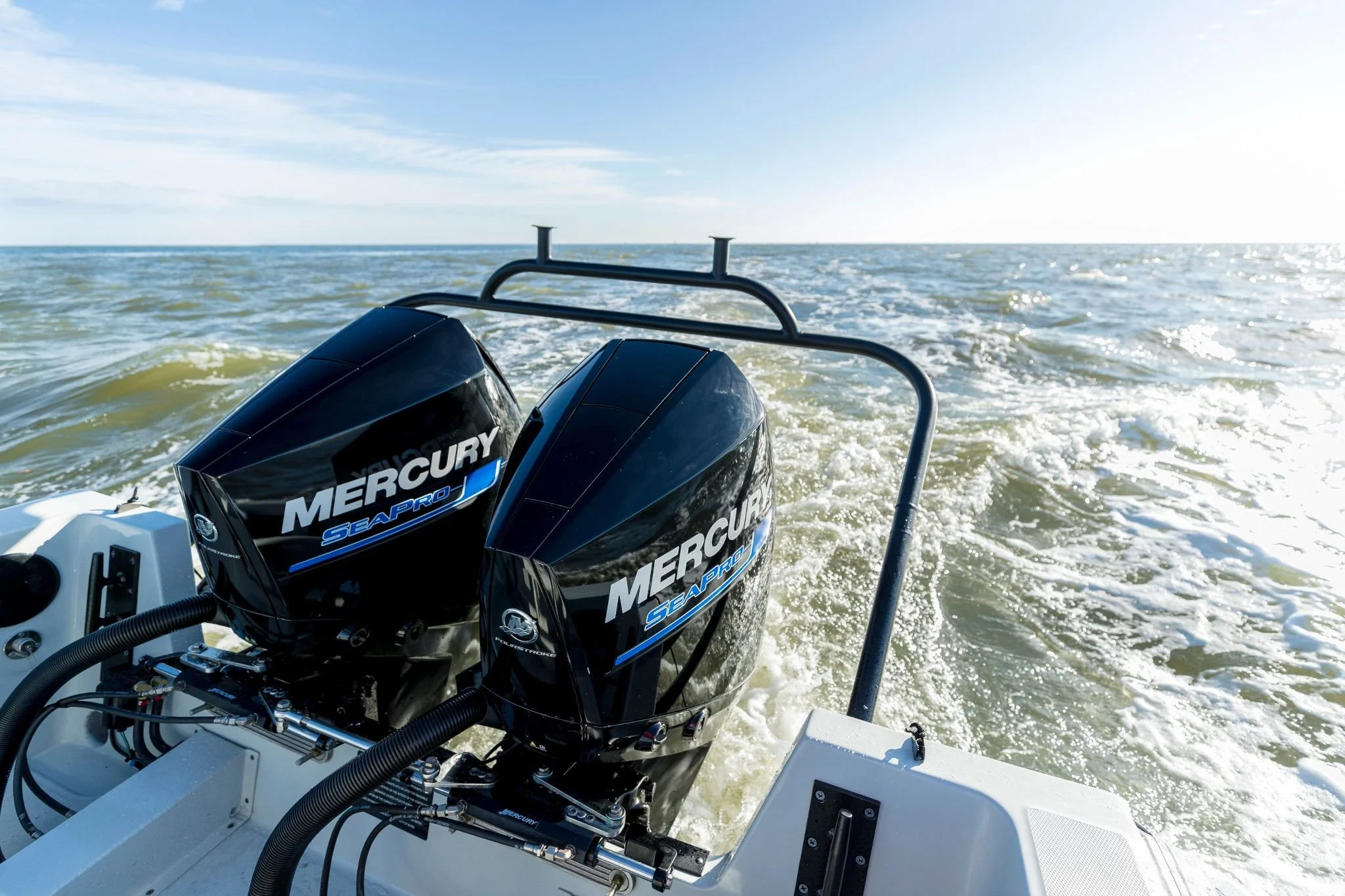 Two Mercury Marine outboard motors on the back of a boat, moving through the water on a sunny day with a clear sky.