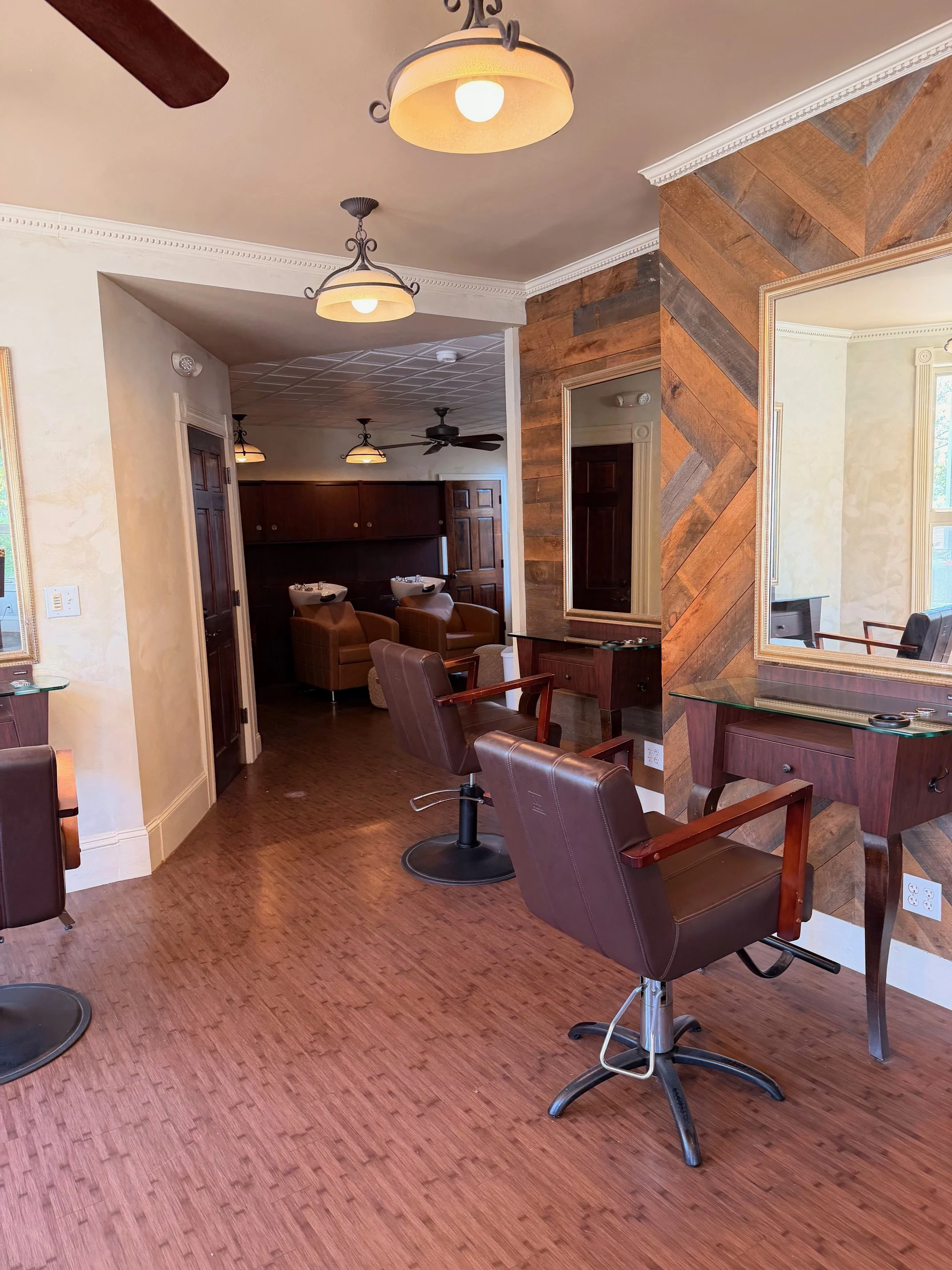 Interior view of a salon with brown leather chairs, wooden flooring, a wooden wall with mirrors, and ceiling lights.