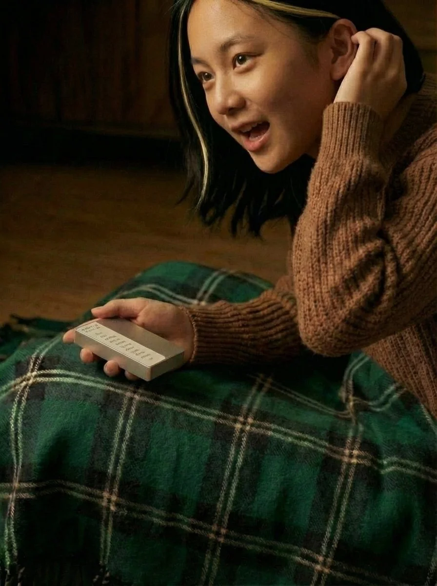 A young woman with long dark hair and a brown sweater sitting on a green plaid blanket, holding a small white box. She looks engaged and happy, possibly talking or laughing.