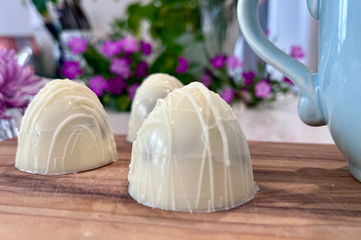 Three white chocolate truffles decorated with white drizzle on a wooden surface with purple and pink flowers blurred in the background.