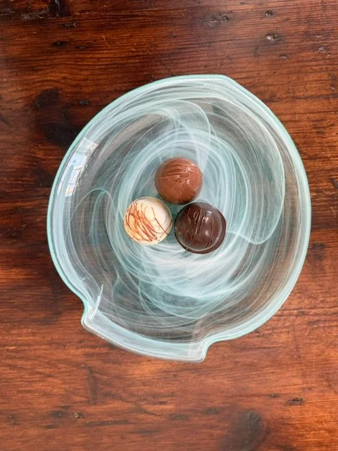 Three assorted chocolate truffles in a glass bowl on a wooden surface.