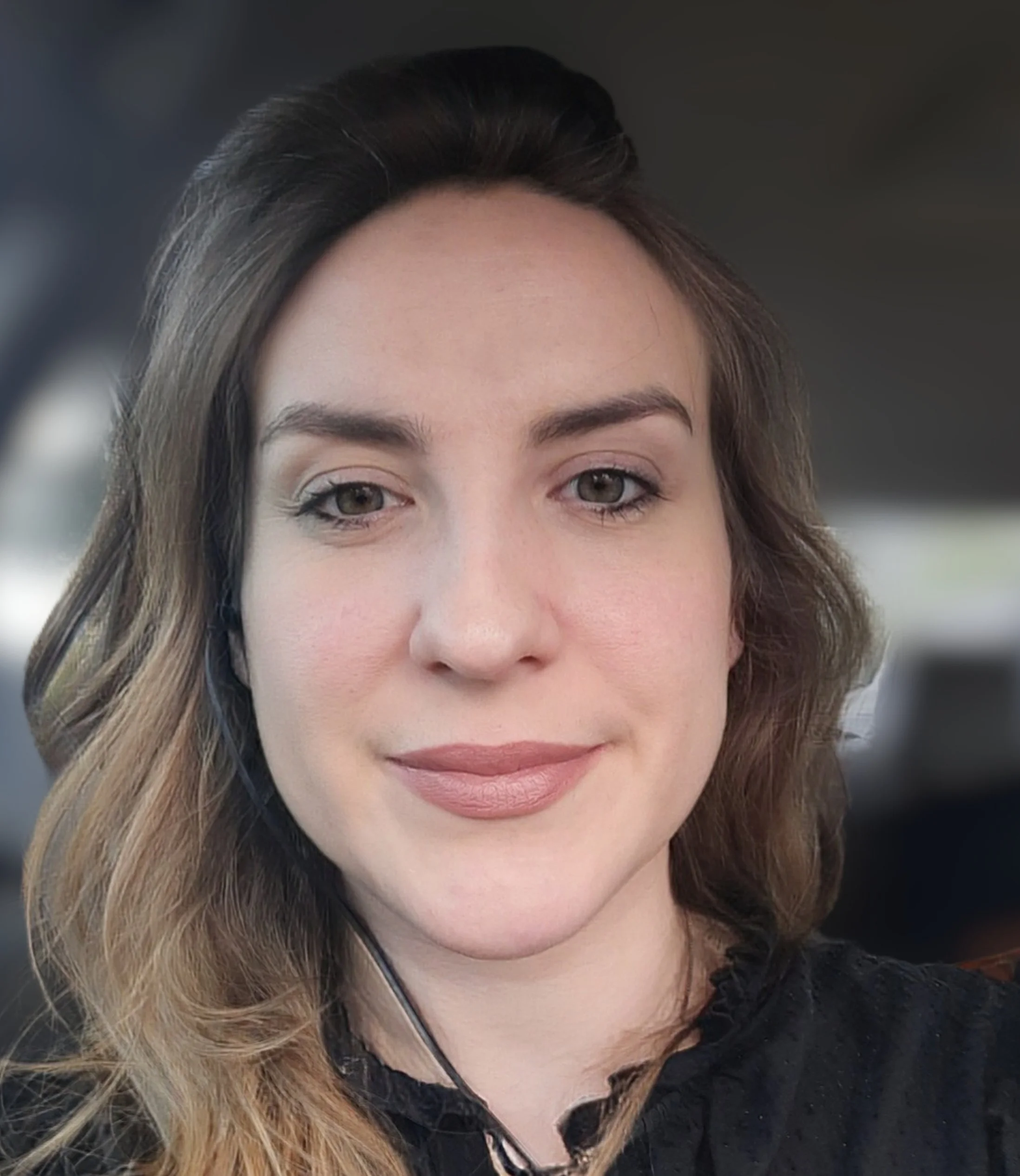 Close-up photo of a young woman with wavy brown hair, light makeup, and a black top, smiling softly inside a vehicle.