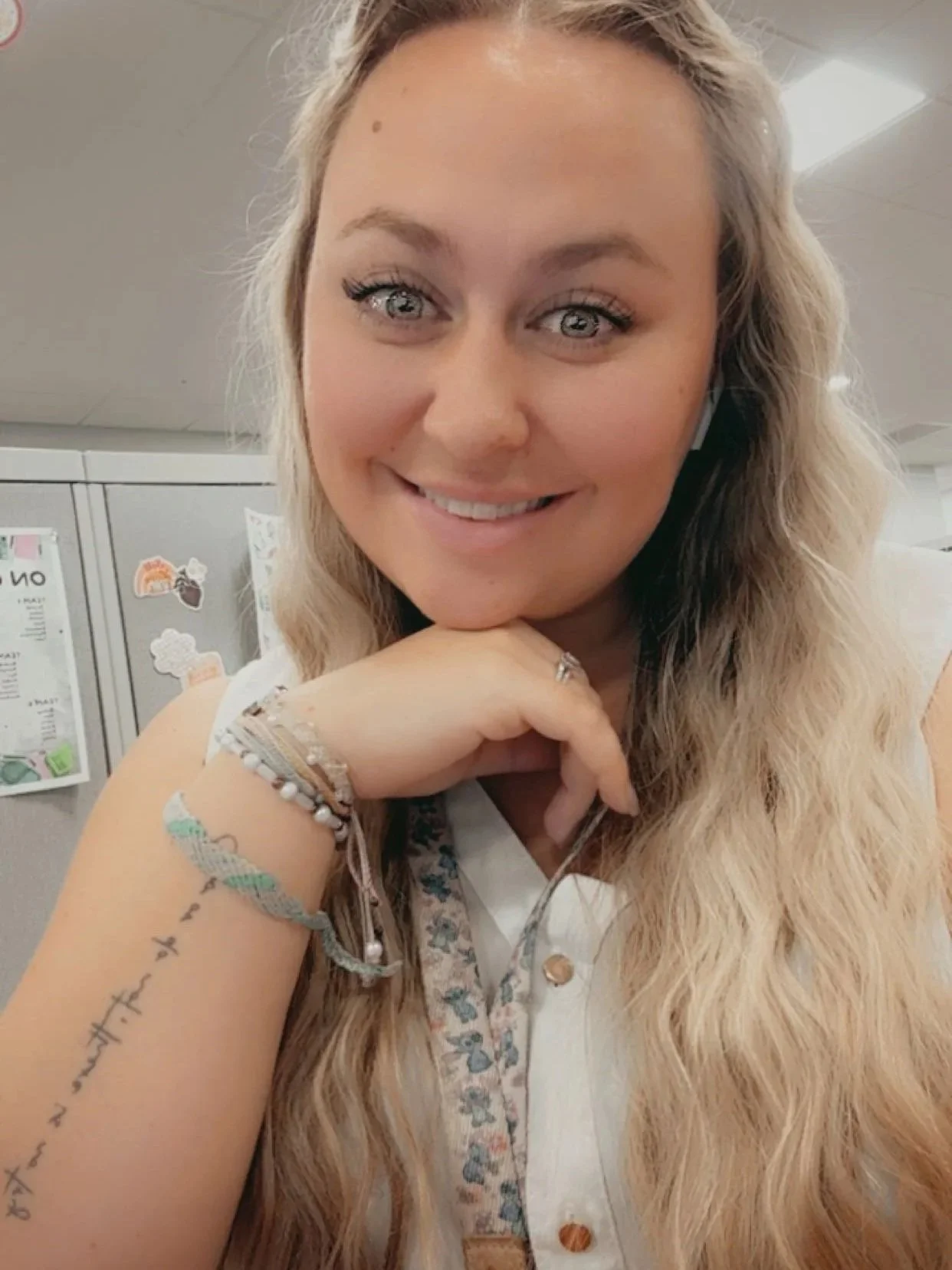 A young woman with long, wavy blonde hair, blue eyes, and a bright smile, resting her chin on her hand in an office cubicle with decorated lockers in the background.