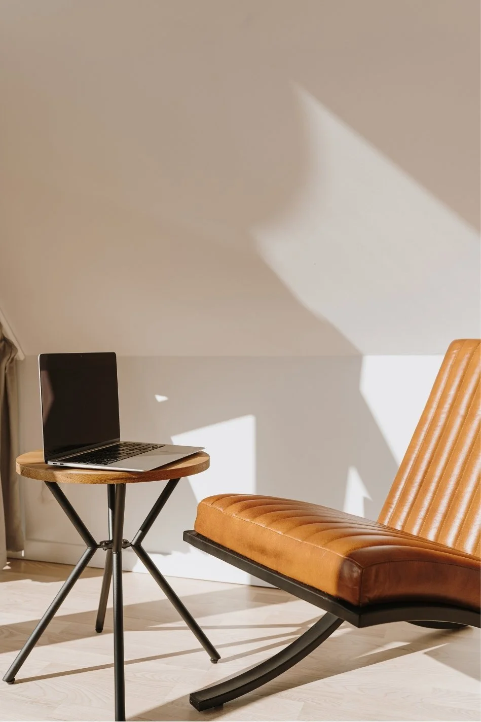 A cozy room with a modern leather lounge chair and a small round wooden table with a laptop on top. Sunlight creates shadows on the wall.