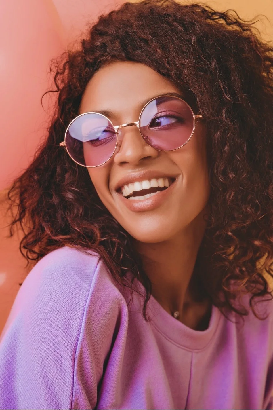 A woman with curly dark hair wearing pink-tinted sunglasses and a light pink shirt, smiling against a warm pink background.