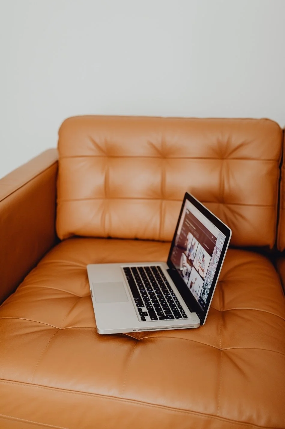 A silver laptop resting on an orange leather couch with tufted backrest and wooden armrest, displaying a webpage with multiple images.
