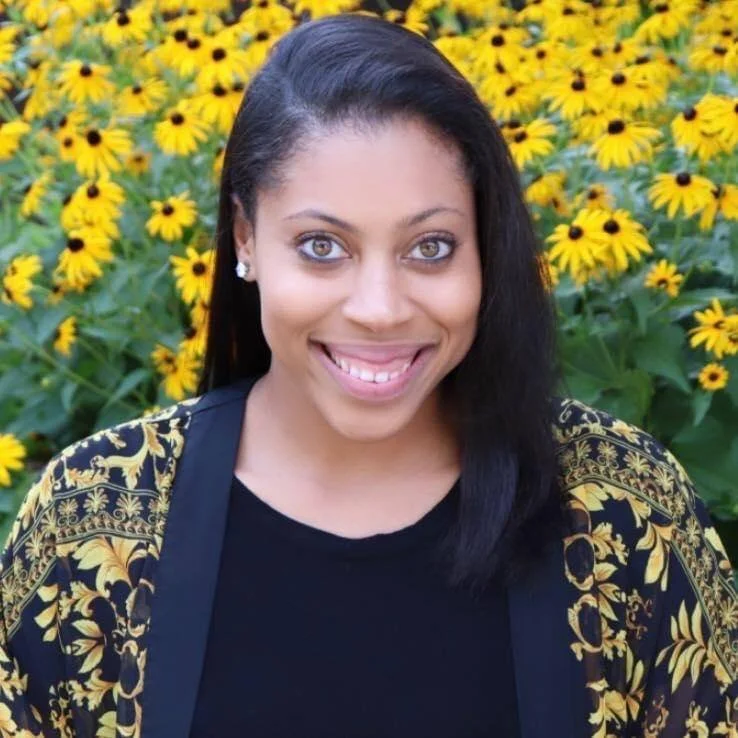 Woman with long black hair smiling, wearing earrings and a black shirt with a patterned black and yellow jacket, standing in front of a field of yellow flowers.