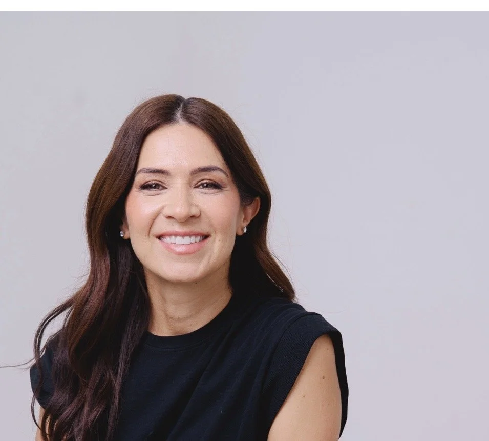 A smiling woman with long brown hair and pearl earrings, wearing a black sleeveless top, against a plain light gray background.
