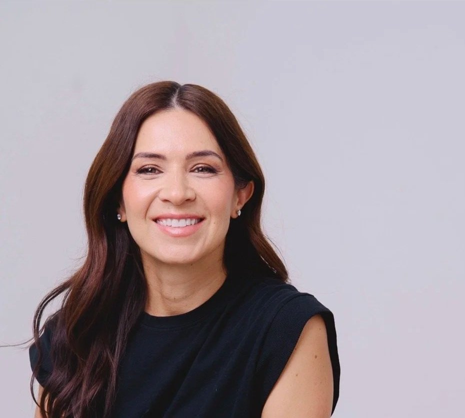 Front view of smiling woman with long brown hair, wearing earrings and a black sleeveless top, against a plain light gray background.