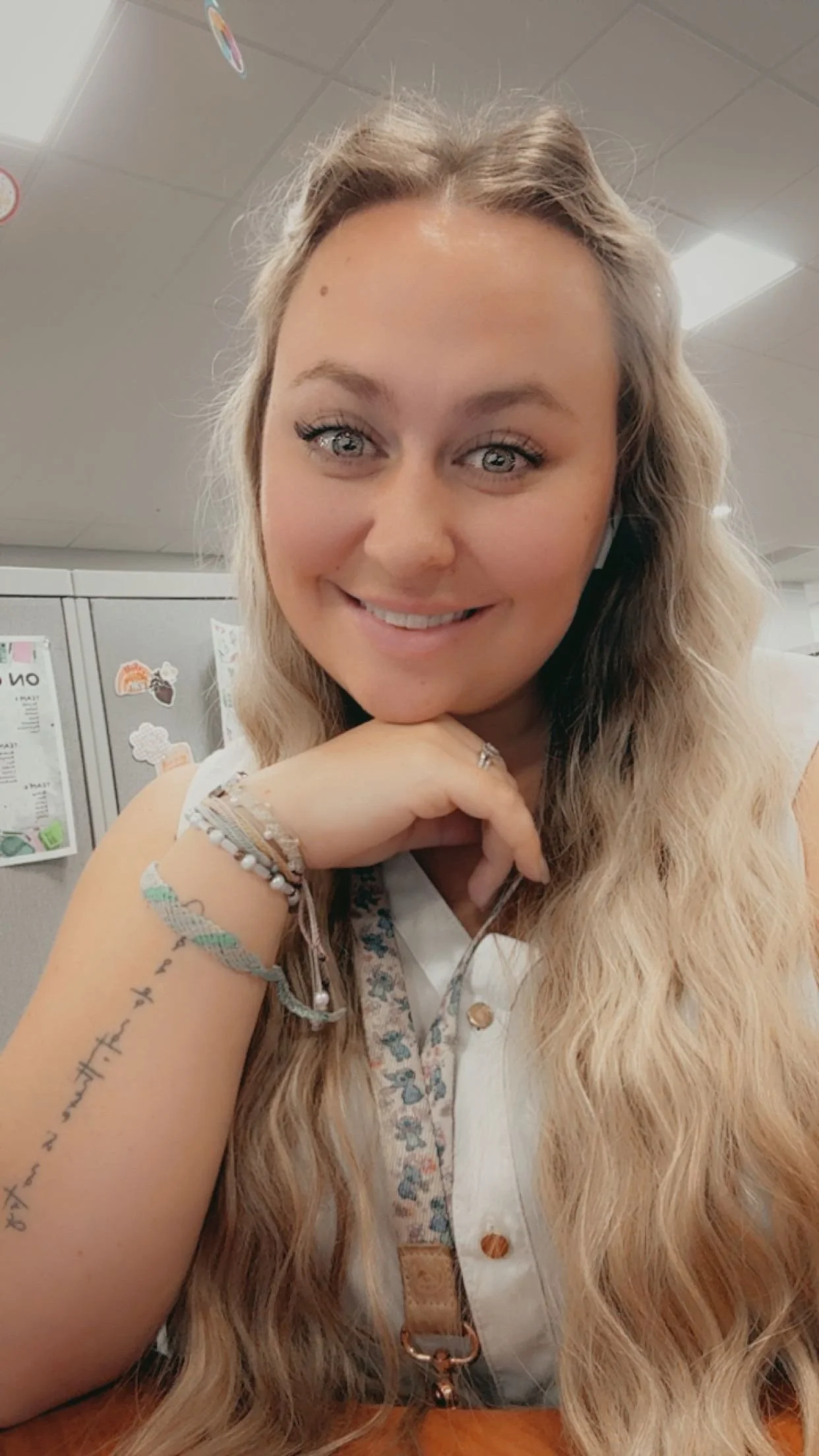 A young woman with long blonde hair smiling at the camera, resting her chin on her hand, in an office environment with lockers and notes in the background.
