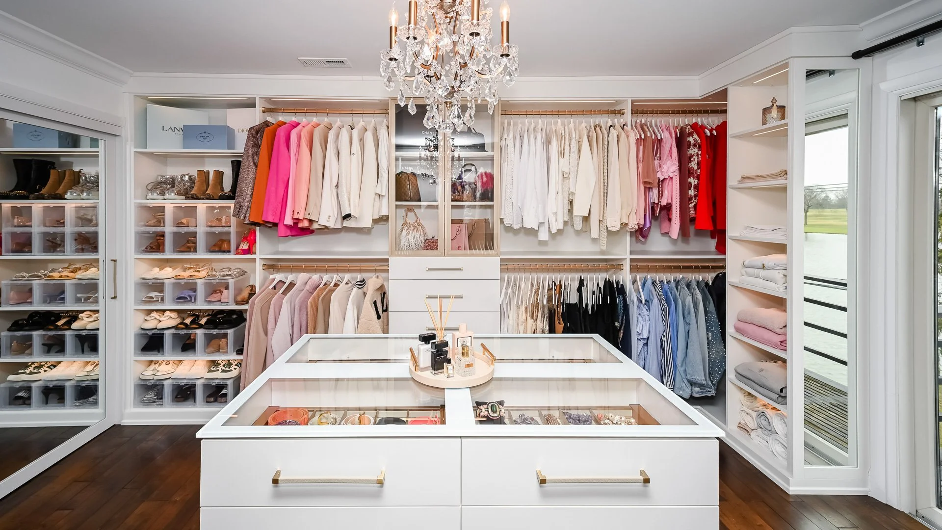 A walk-in closet with white shelves and hanging clothing, including blouses, jackets, and dresses in various colors. A glass-topped jewelry display table is in the center, and a chandelier hangs from the ceiling. The room has a sliding glass door on 