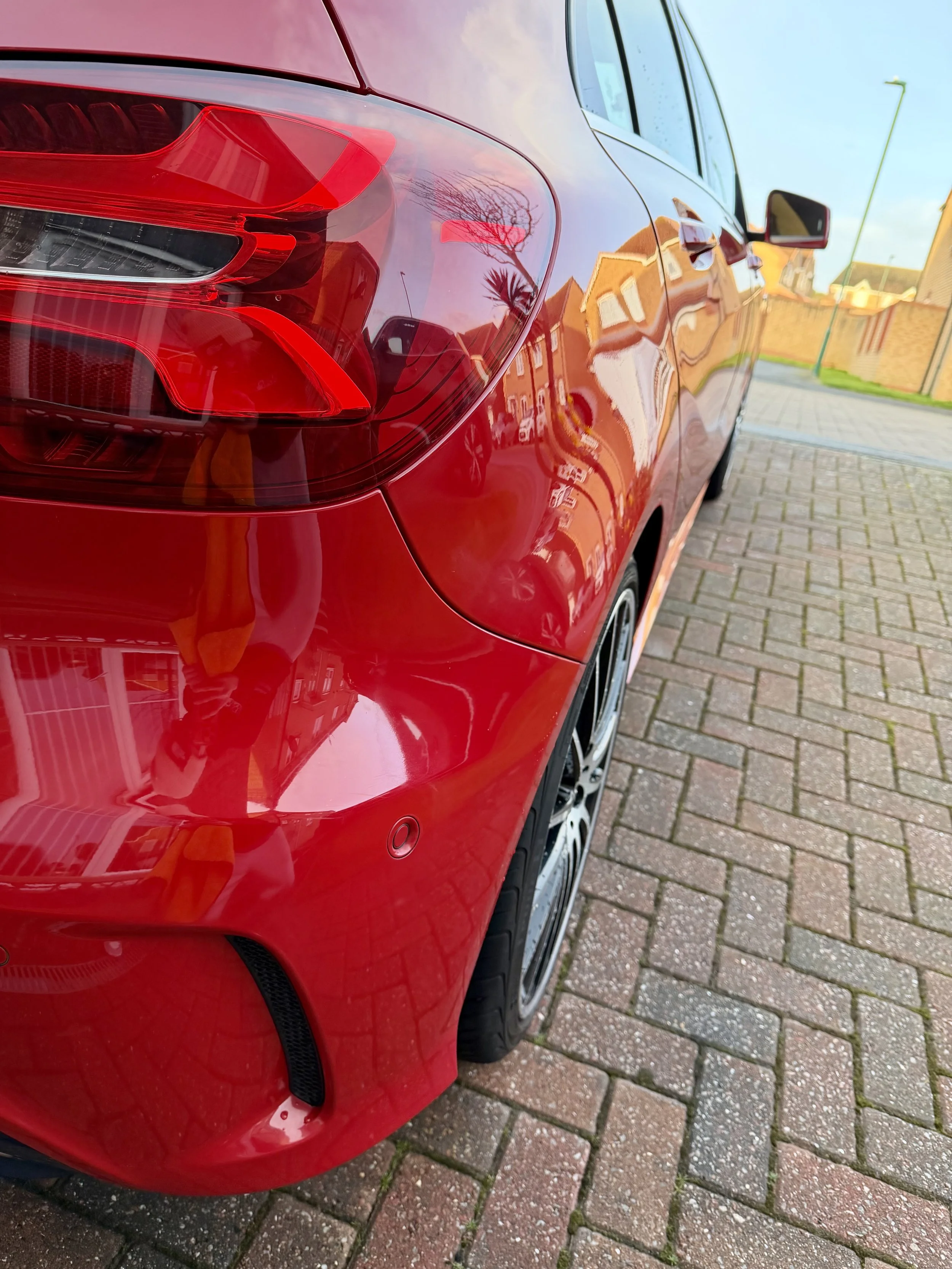 Close-up of the rear left side of a shiny red car parked on a brick driveway, showing the tail light, part of the rear bumper, and the wheel.