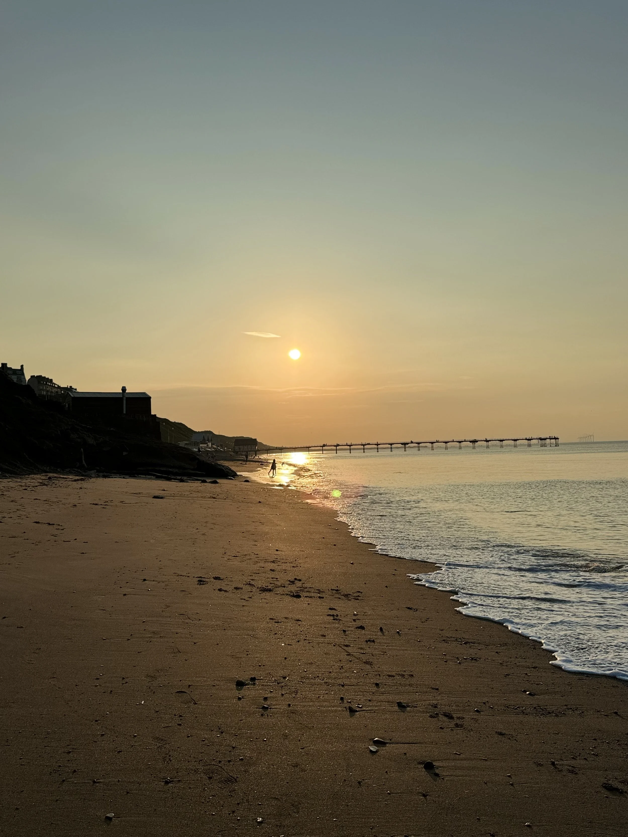 Sunset over a sandy beach with a pier in the distance and a person walking near the shoreline.