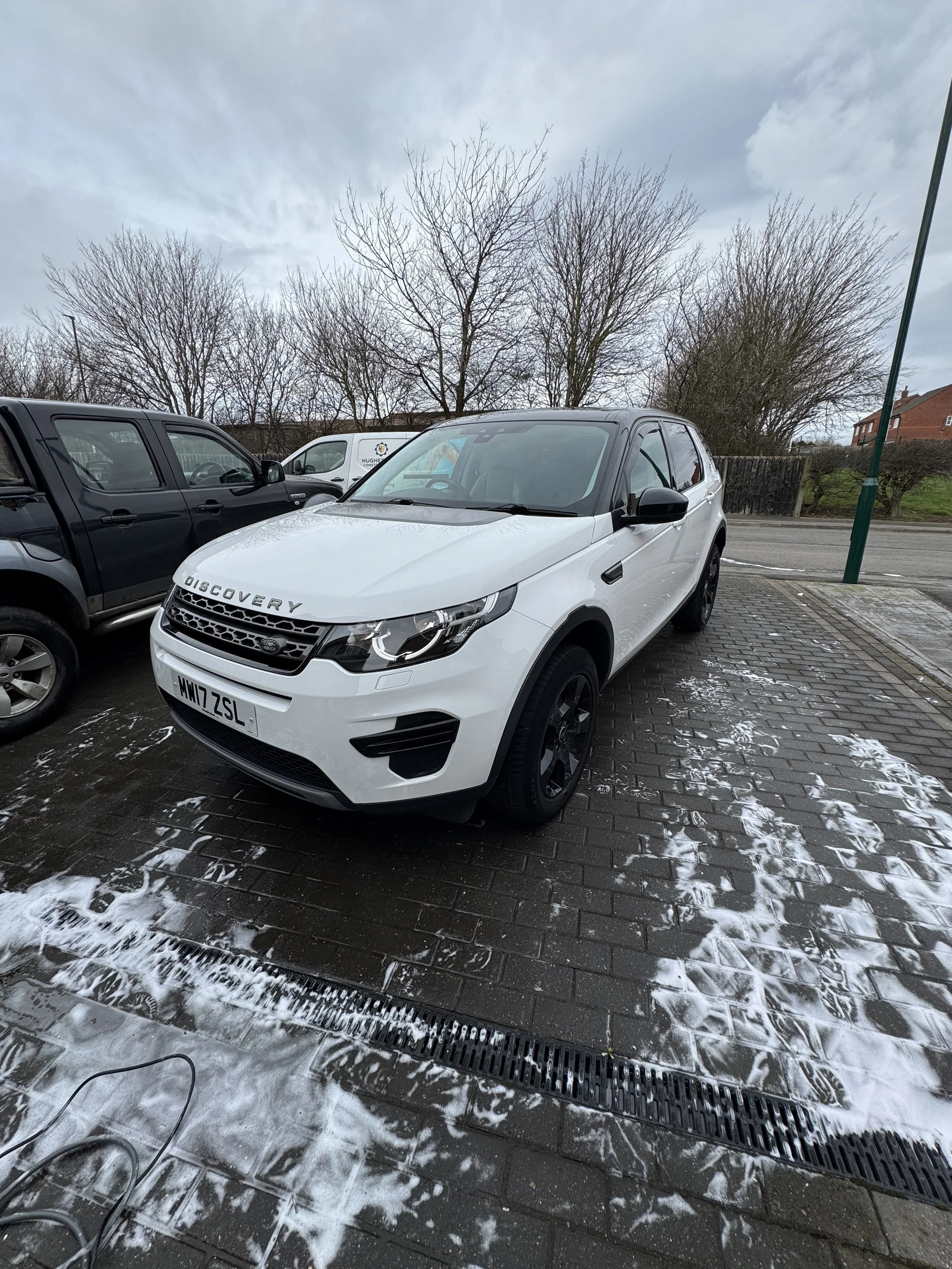 White Land Rover Discovery parked on a brick surface with soap suds, surrounded by other vehicles and leafless trees in the background.