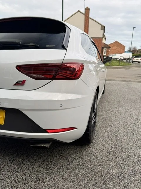 White hatchback car parked in a residential area with houses and other parked cars visible in the background.