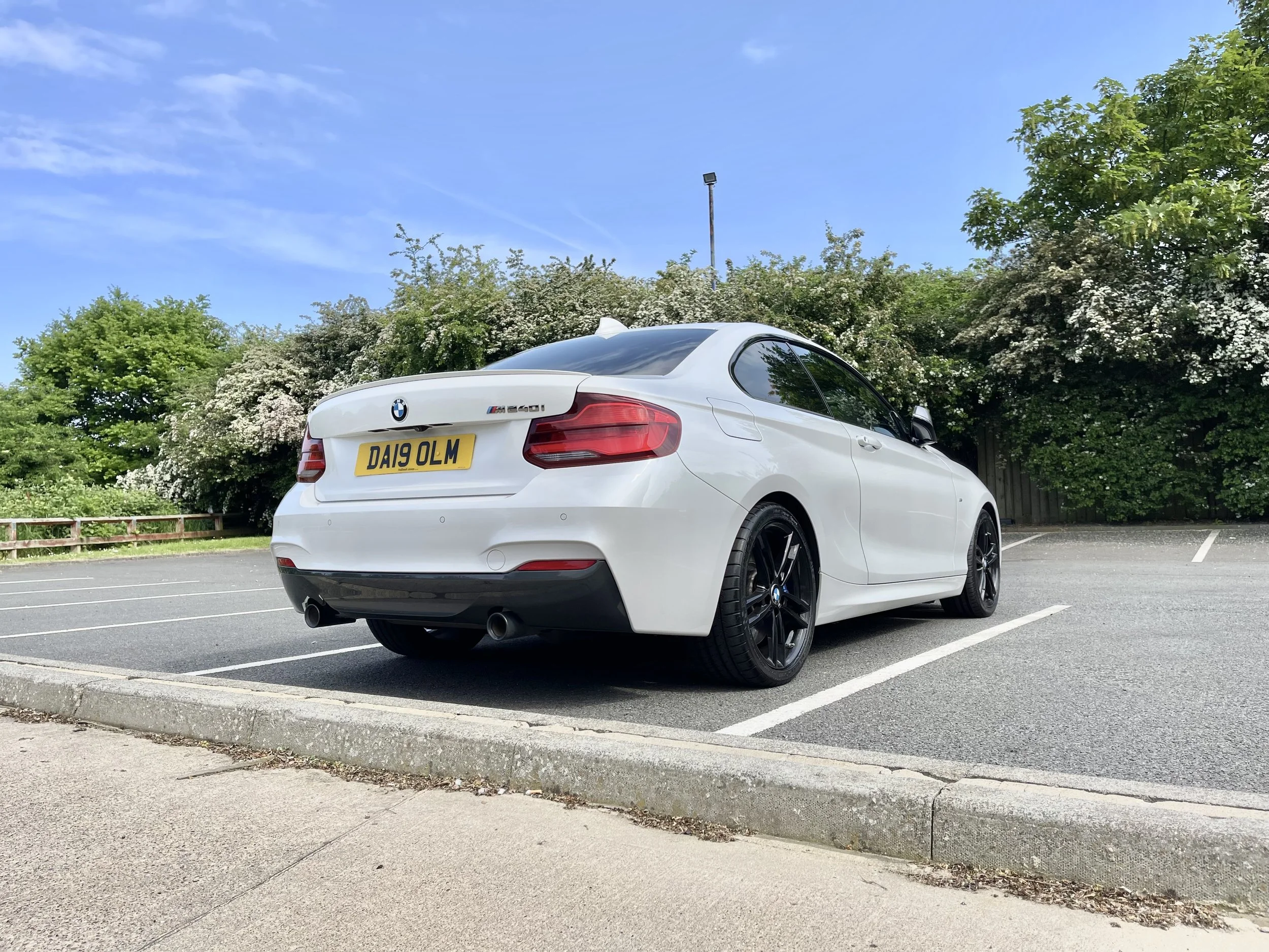 White BMW convertible sports car parked in an empty parking lot on a sunny day with trees and blue sky in the background.