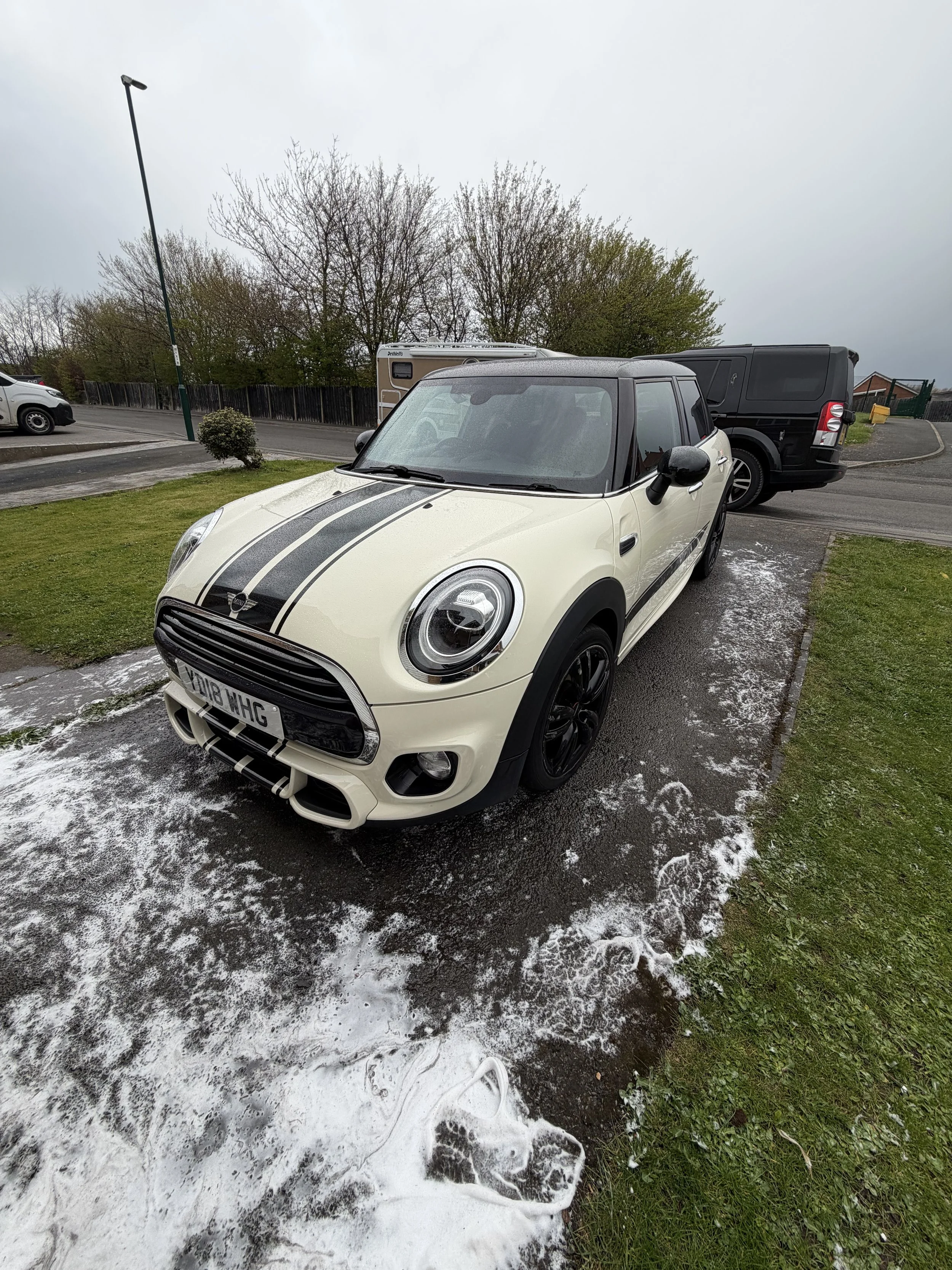 A white Mini Cooper with black racing stripe parked on a wet outdoor surface, surrounded by patches of foam or soap, with other vehicles and trees in the background.