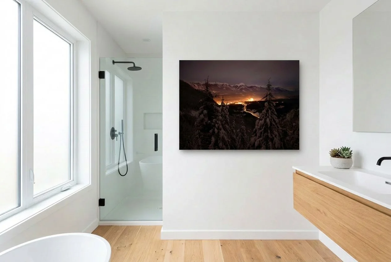 Modern bathroom with white walls, a large window, a walk-in shower, a wall-mounted television displaying a mountain landscape at sunset, a wooden vanity with a white countertop, and a small potted succulent plant.