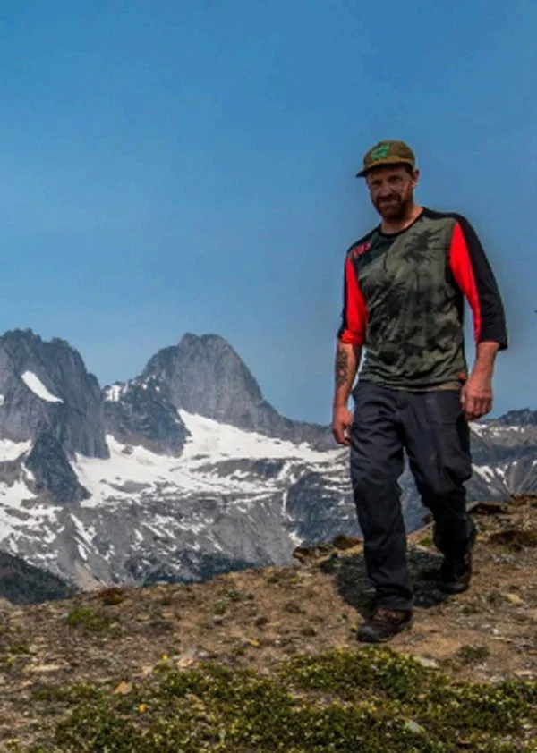 A man with a beard in outdoor gear standing on rocky ground at a mountain landscape with snow-covered peaks and a clear blue sky.