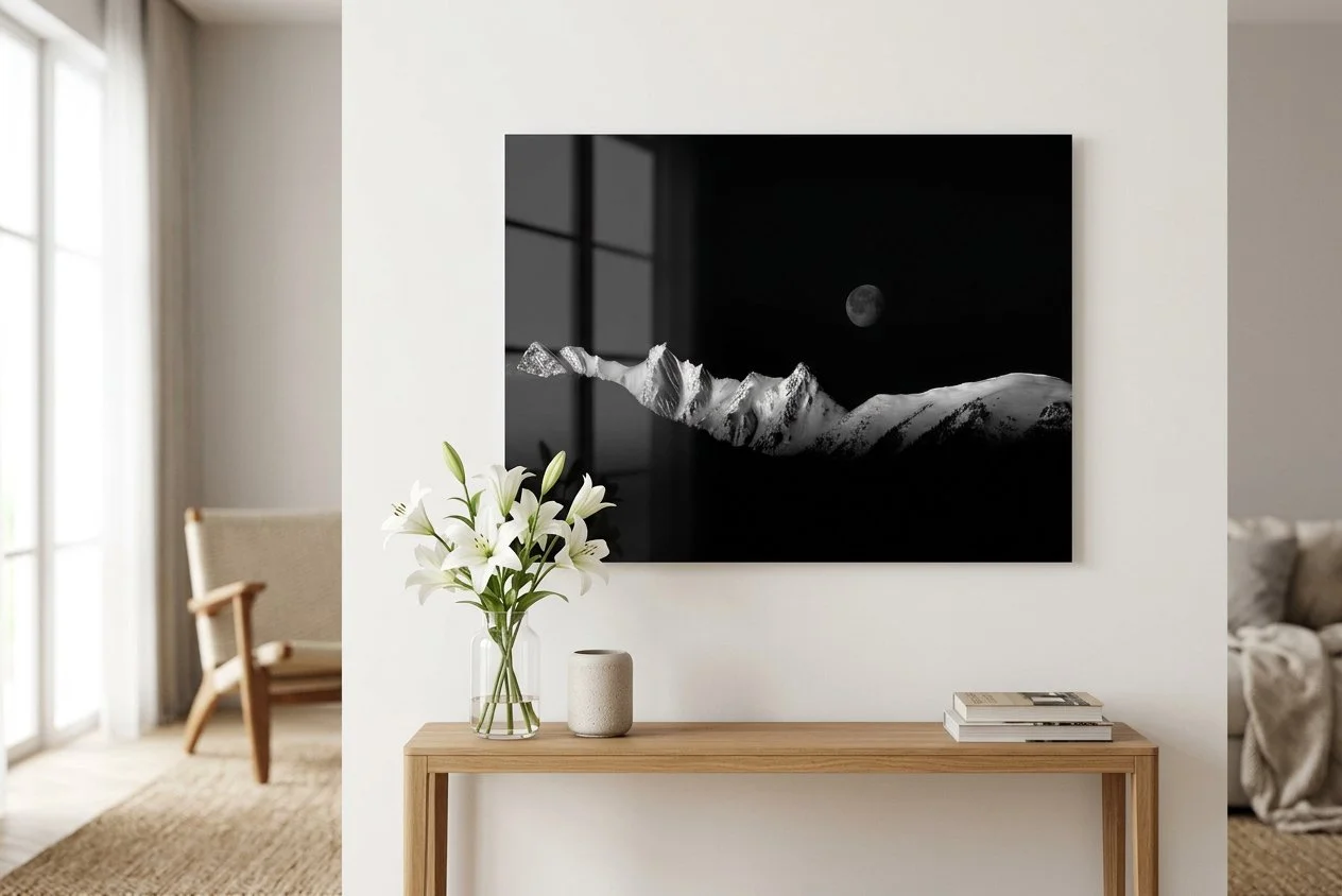Modern living room with a wooden console table, a vase of white lilies, a small beige decorative object, and stacked books. A large framed black-and-white photograph of snowy mountains and the moon hangs on the white wall. A beige armchair and a window with curtains are visible in the background.