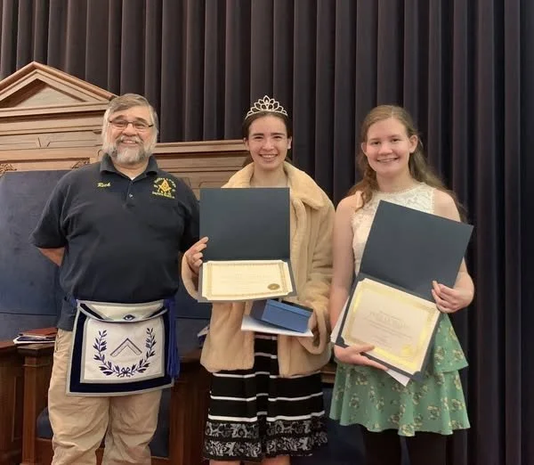 Two young girls holding award certificates and a man standing next to them, all smiling, in a formal setting with dark curtains and wood paneling in the background.