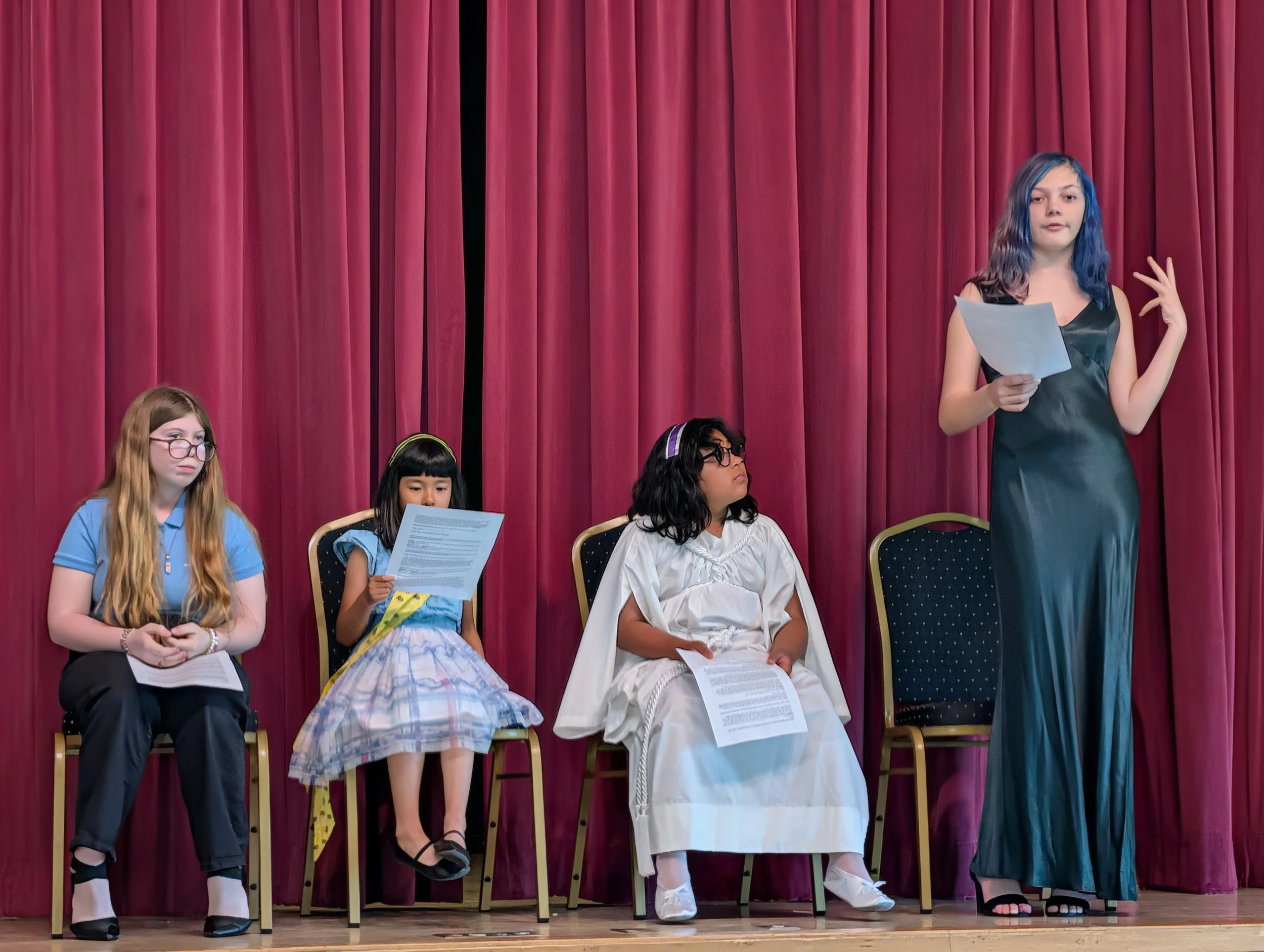 Several children sitting on chairs on stage with a red curtain behind them. One girl standing and speaking, holding a paper.