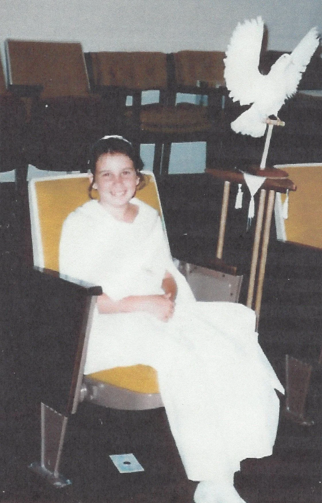 A young girl dressed in a white robe, sitting in a yellow and black chair in a room with dark wood chairs and a table with a white cloth. There is a large white dove-shaped cutout on a stick in the background.