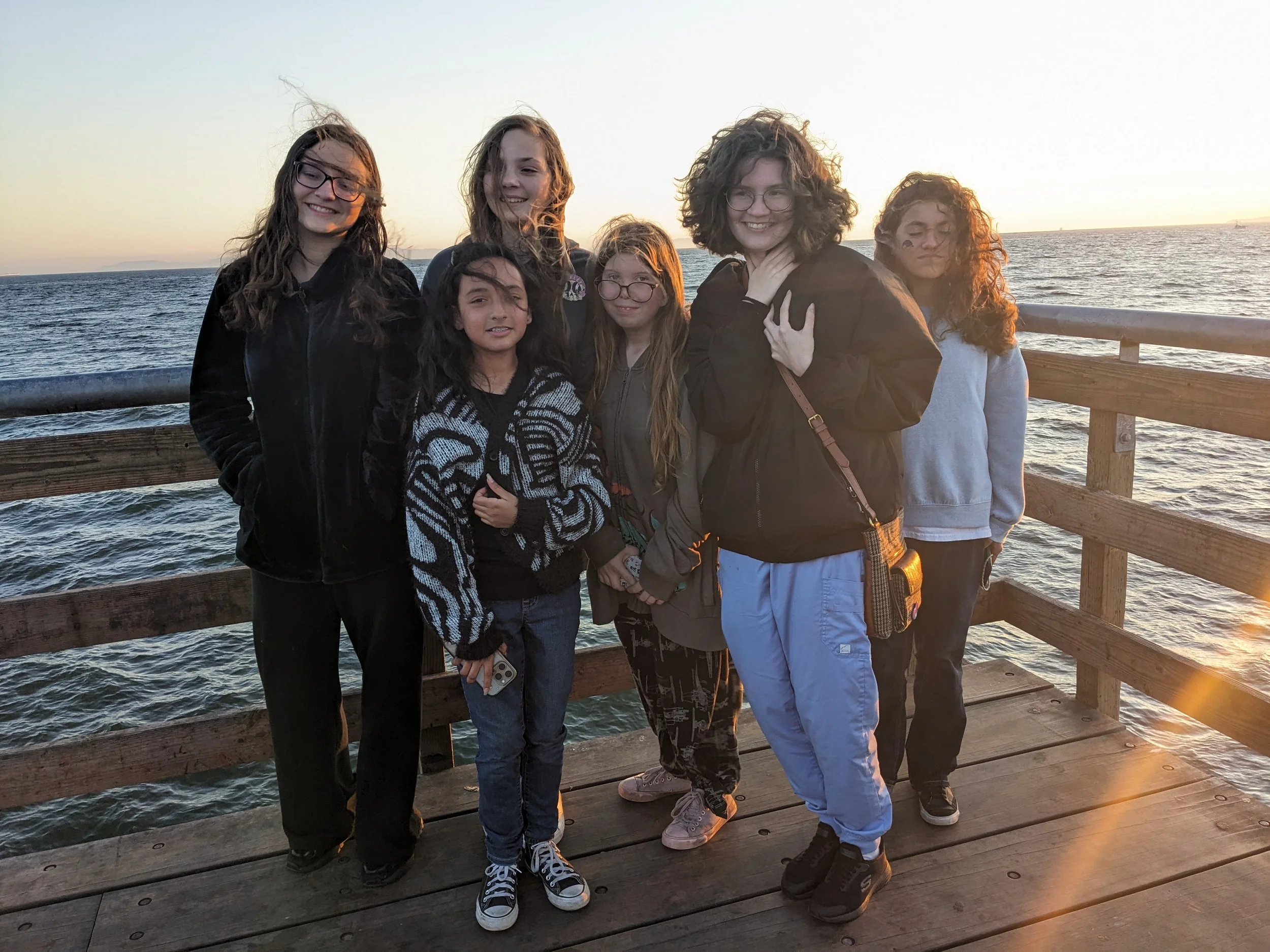 Seven young girls standing on a wooden pier by the water at sunset, smiling and posing for the photo.