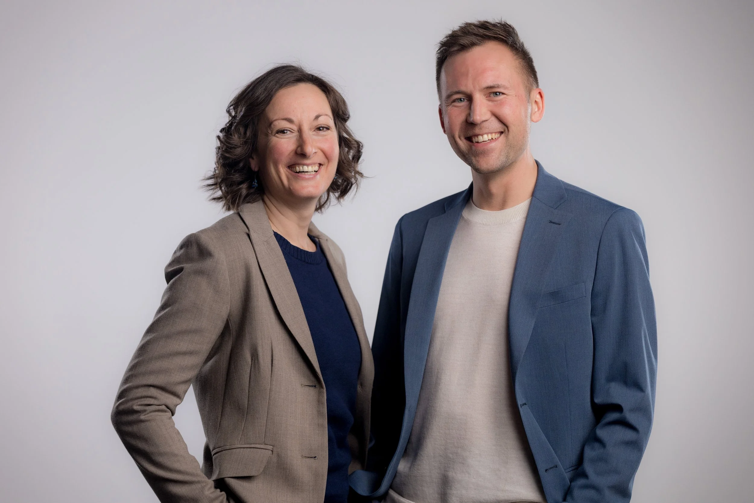 Two smiling professionals, a woman and a man, standing side by side against a plain gray background, dressed in business casual attire.
