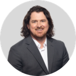 A man with shoulder-length dark hair and a beard, wearing a dark suit and white shirt, smiling, against a grey circular background.