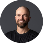 Portrait of a man with a beard and shaved head smiling against a dark background.