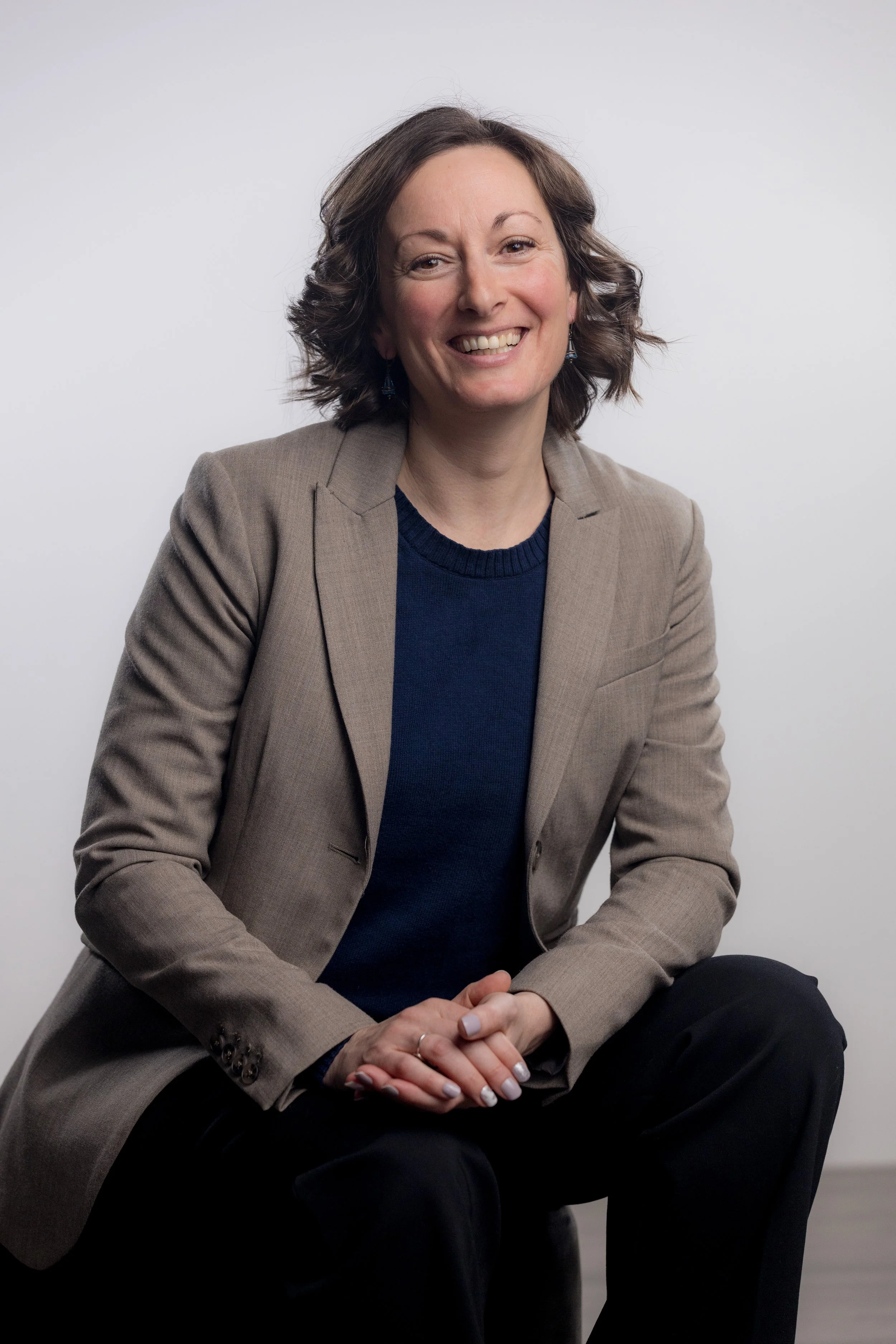 A woman with curly brown hair, wearing a beige blazer over a navy blue top, smiling, sitting with her hands clasped on her lap against a plain white background.