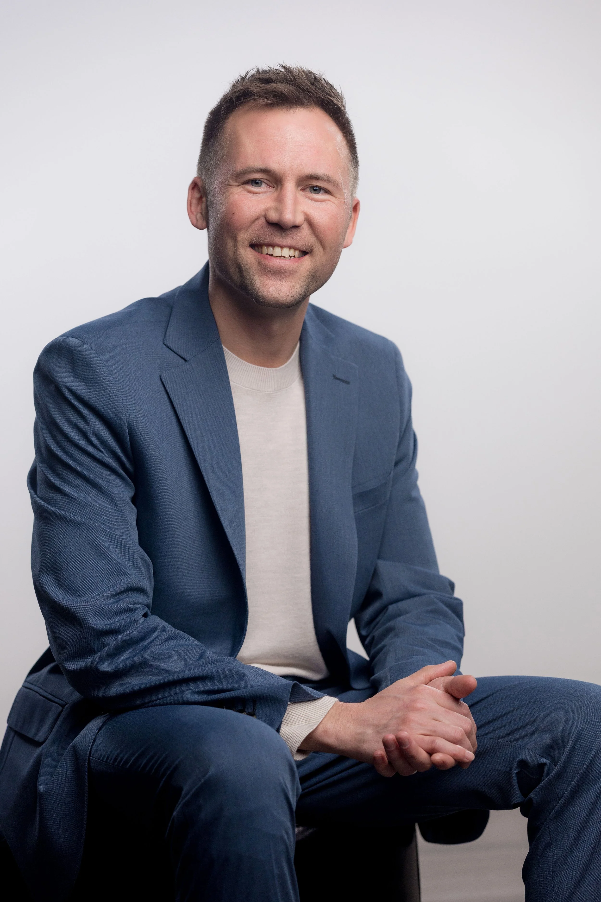 A smiling man in a blue blazer and white shirt sitting with hands clasped in front of him, against a plain white background.