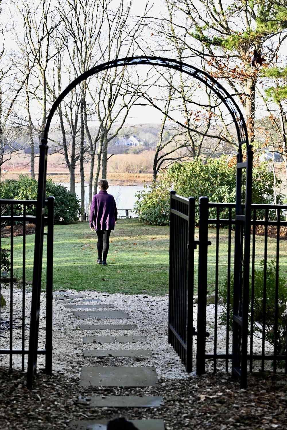 Person walking through an open garden gate toward a peaceful lake, symbolizing a new beginning on the spiritual journey