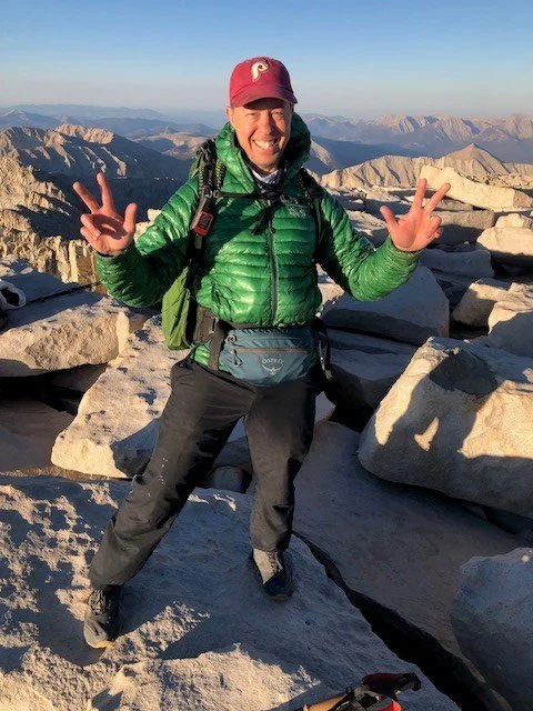 A man standing on rocky terrain in a mountain landscape, smiling, making peace signs with both hands, dressed in outdoor hiking gear including a green jacket, red cap, and backpack.