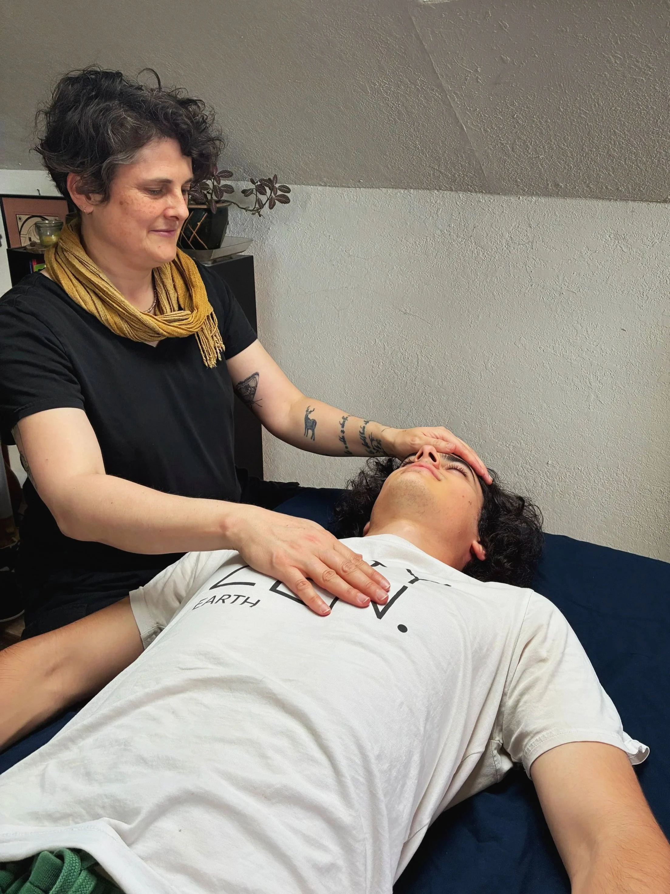 A woman with curly dark hair giving a massage to a man lying on a massage table. The woman has tattoos on her left arm and is wearing a black shirt and a yellow scarf. The man has dark curly hair and is wearing a white T-shirt.