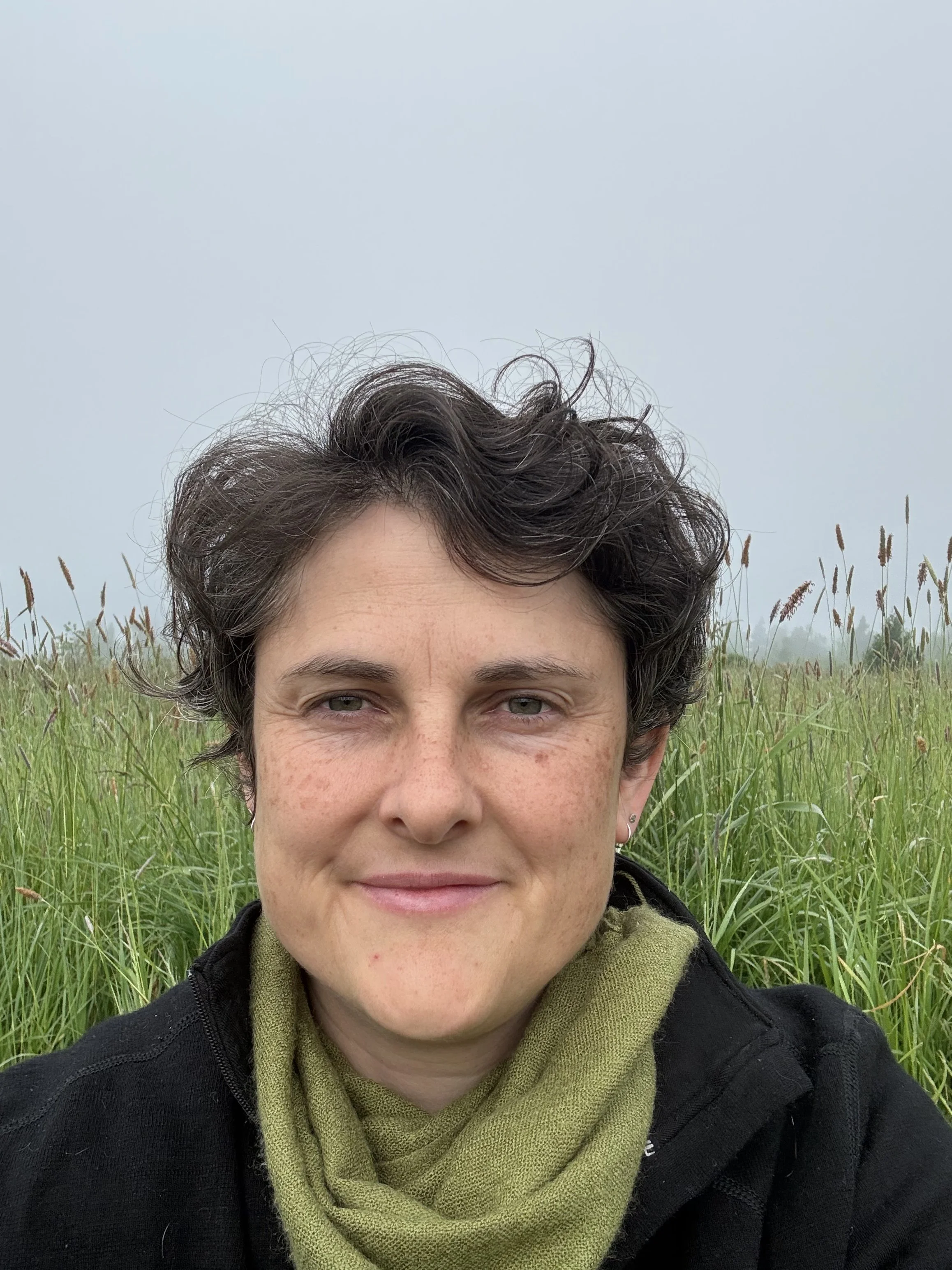 A woman with short curly dark hair and light skin taking a selfie outdoors in a grassy field with tall grass and a cloudy sky in the background.
