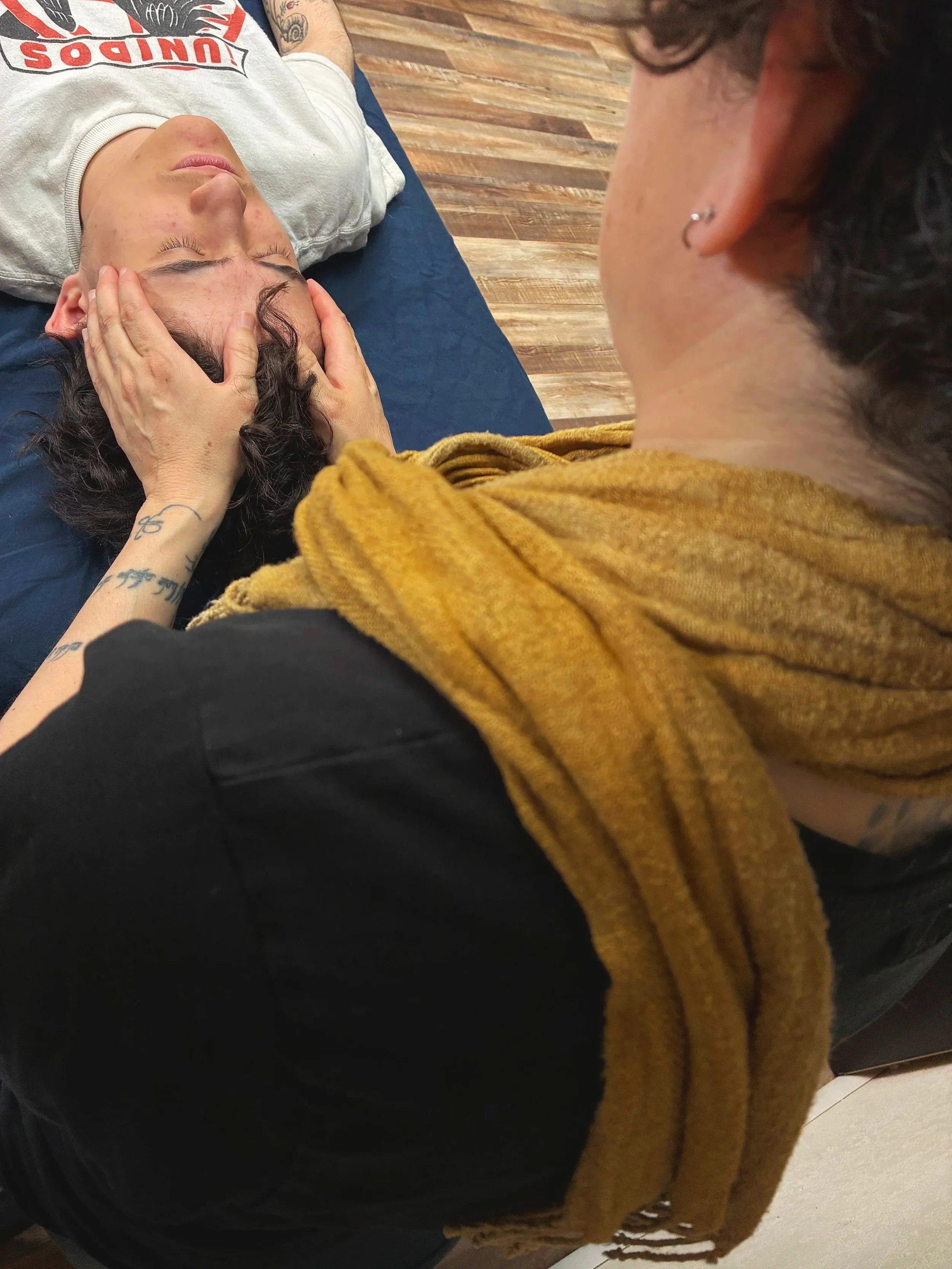 A person lying on their back with hands cradling their head, eyes closed, and a second person leaning over them. They are indoors on a wooden floor.