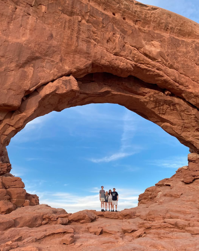 Three people standing under a large red rock arch in a desert landscape with a bright blue sky and wispy clouds