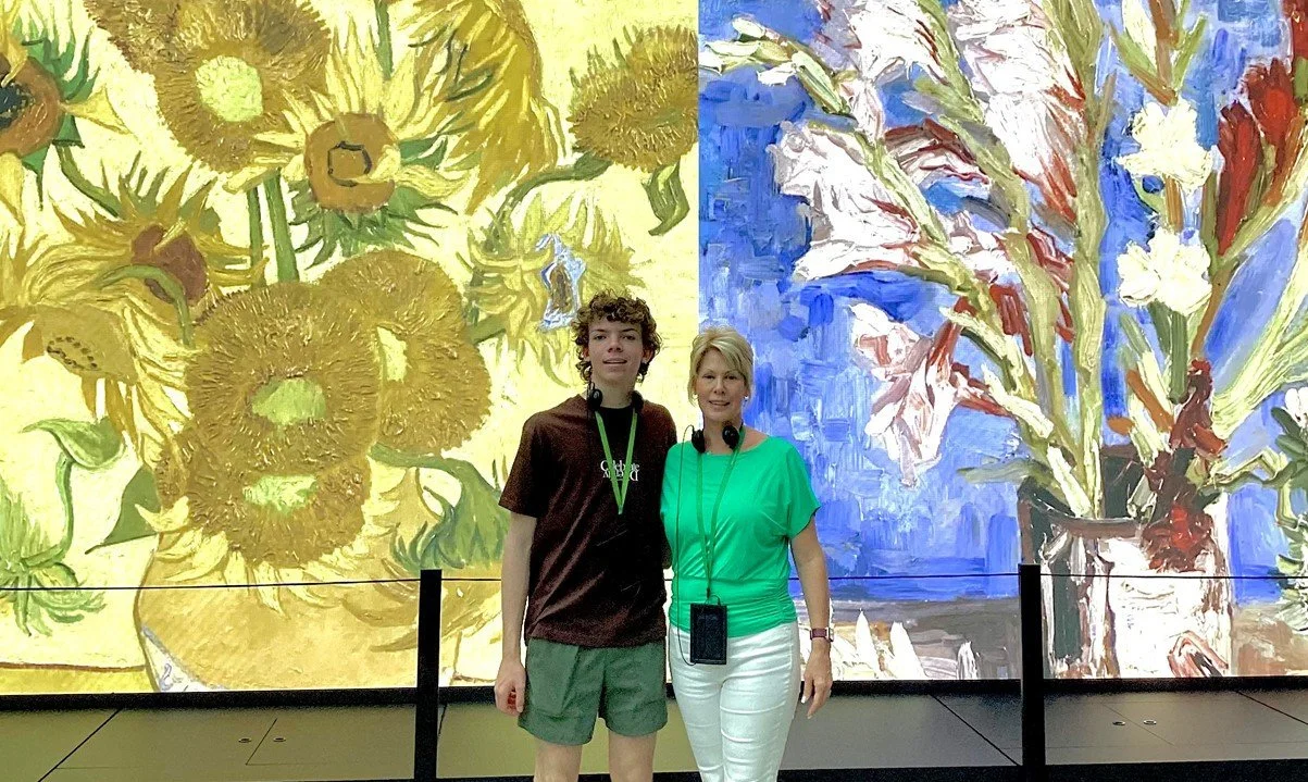 Two people standing in front of large digital screens displaying paintings of sunflower and iris flower arrangements, one woman and one young man.