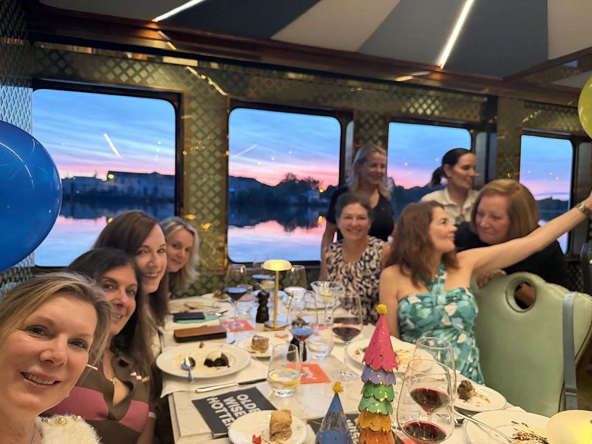 A group of women celebrating at a dinner party on a boat with a sunset view through the windows, decorated with balloons and party hats, enjoying food and drinks.