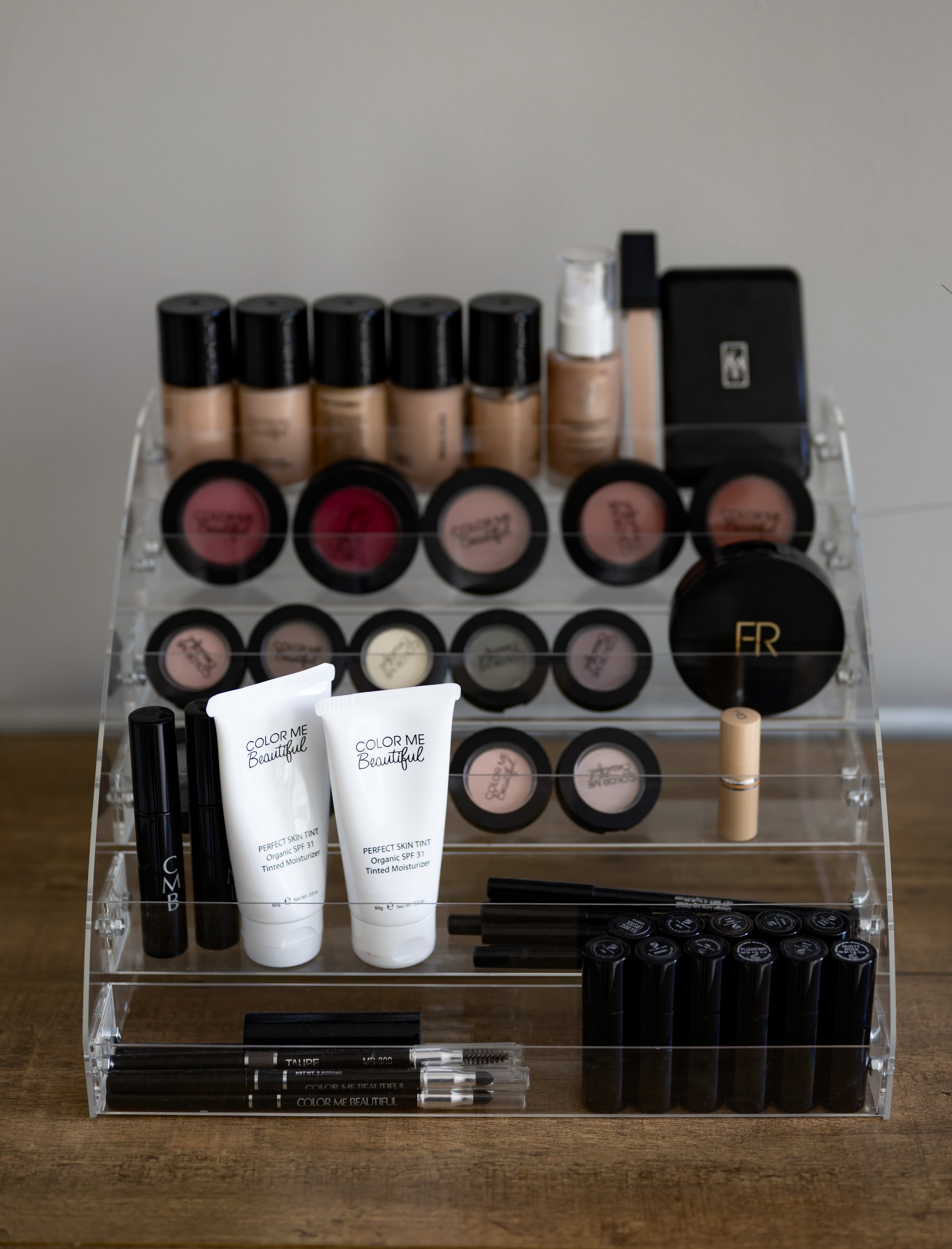 A clear acrylic makeup organizer with multiple tiers, holding foundation bottles, lip glosses, a brow pencil, and other cosmetic products on a wooden surface.