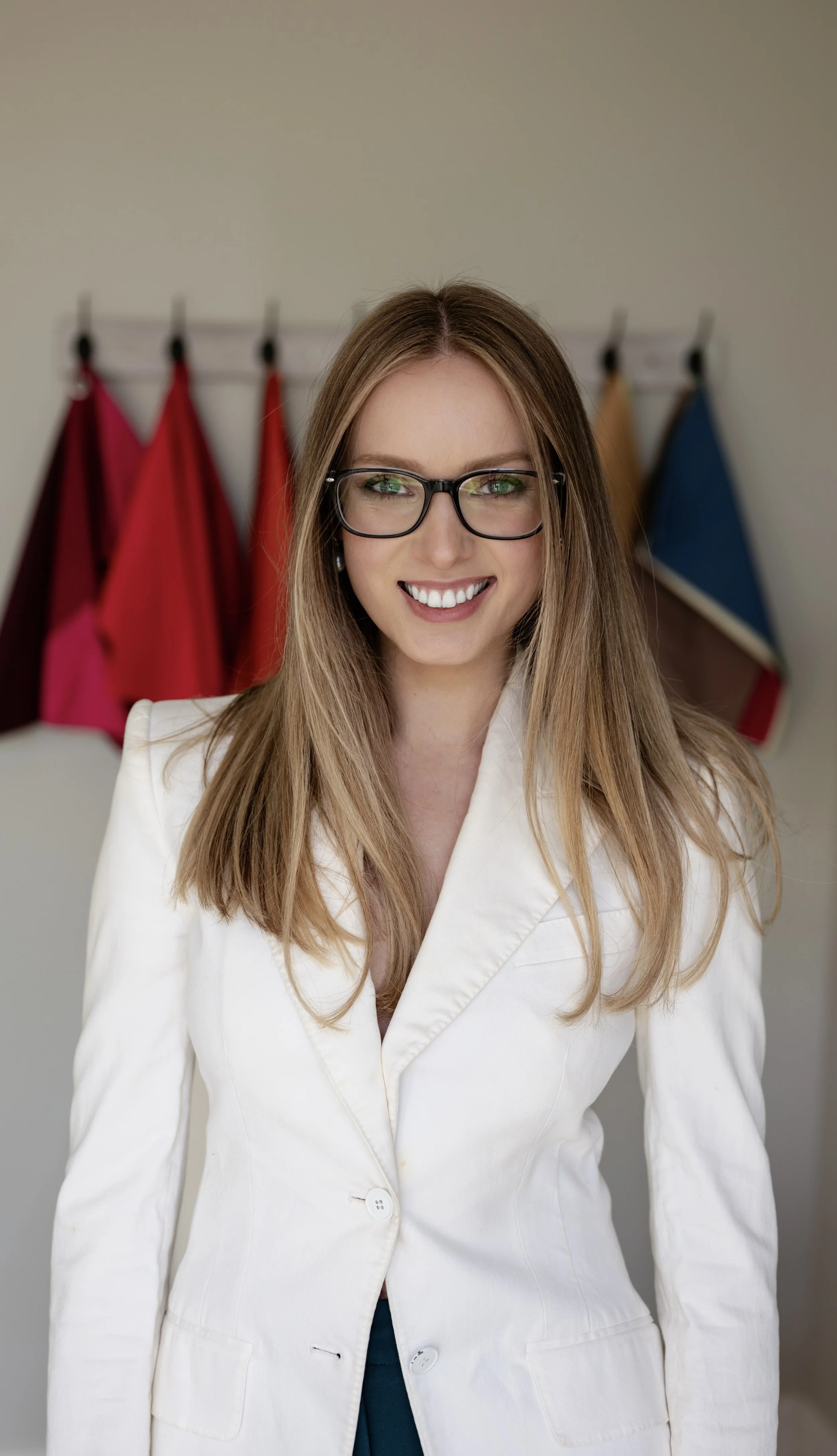 A woman with long light brown hair wearing glasses and a white blazer, smiling in front of a background with colorful flags.