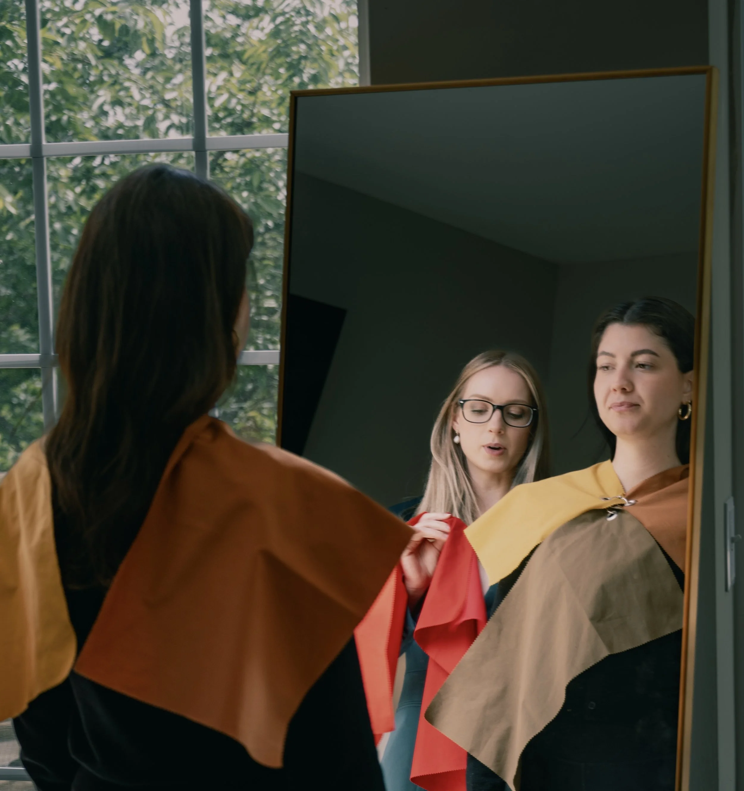 People standing in front of a mirror, with windows showing green trees outside, reflecting a conversation or preparation.